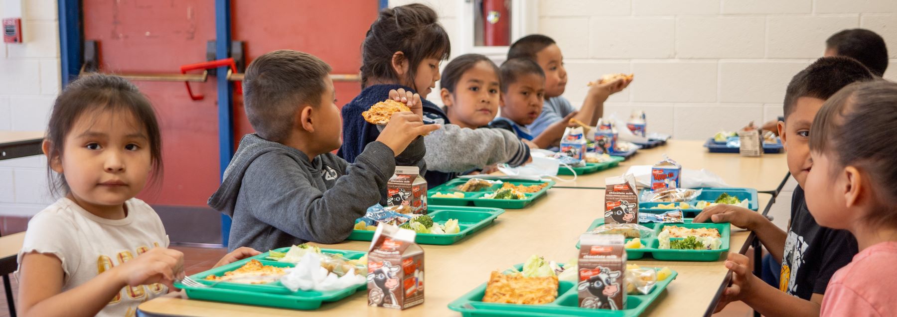 A group of children seated at a lunch room table, happily eating lunch and engaging in conversation with one another.