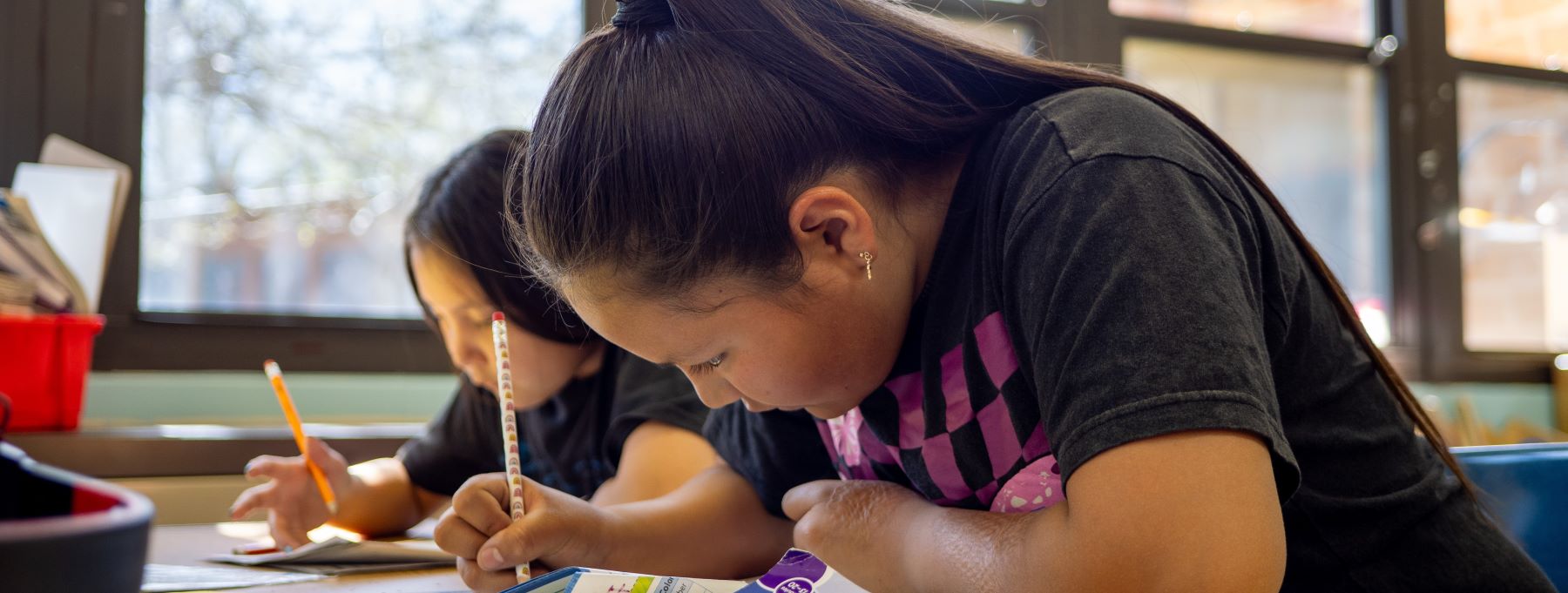Two students sit at a desk in a classroom, focused on their work with books and papers spread out in front of them.
